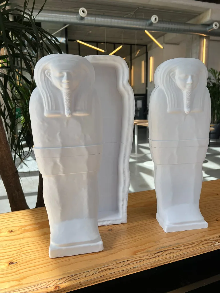 White 3D printed Egyptian sarcophagus prototypes displayed on a wooden table in the MadeInBXL Brussels workshop